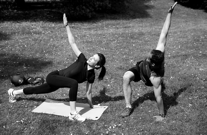 Roland and a client doing a side-plank twist on mats in the park, in black and white.