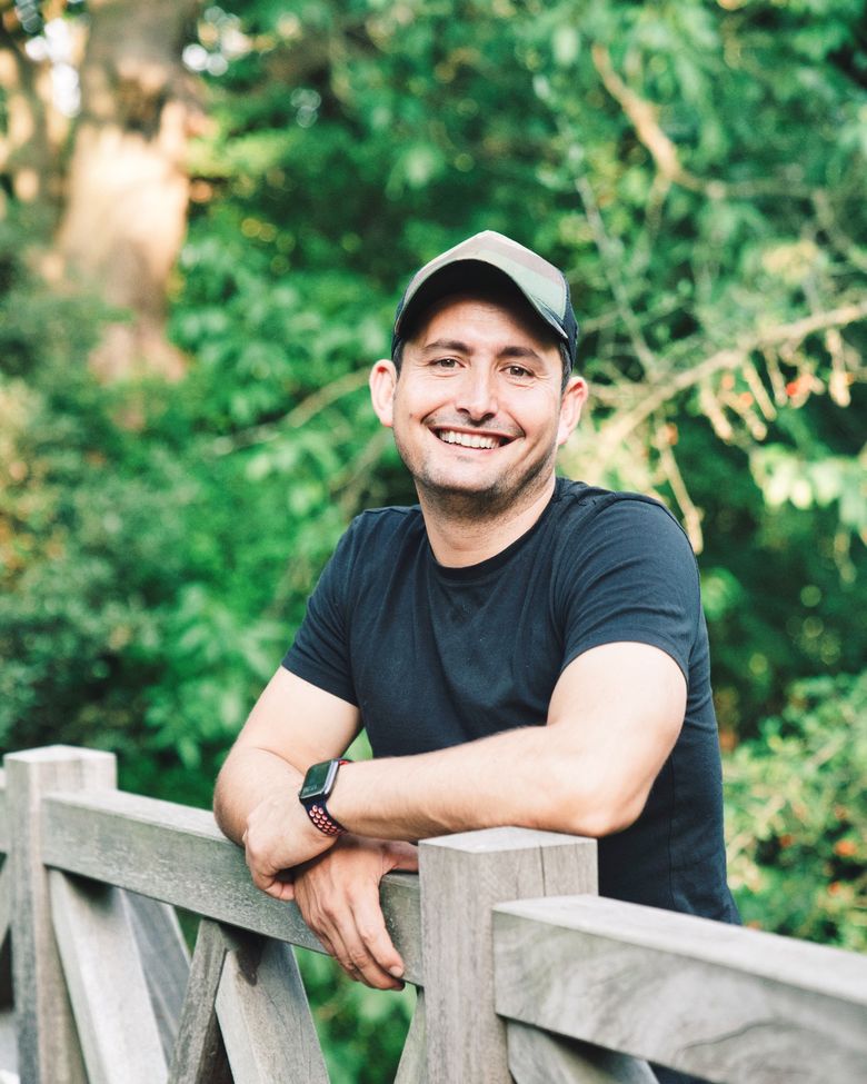 Roland Feizo smiling, in a cap and black T-shirt, in front of green trees.