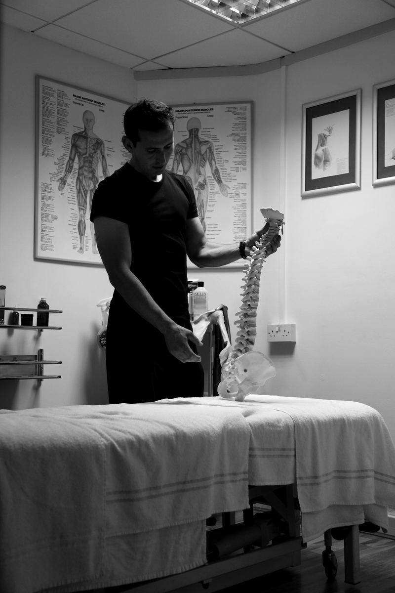 Roland holding a model spine beside a massage couch, in black and white.